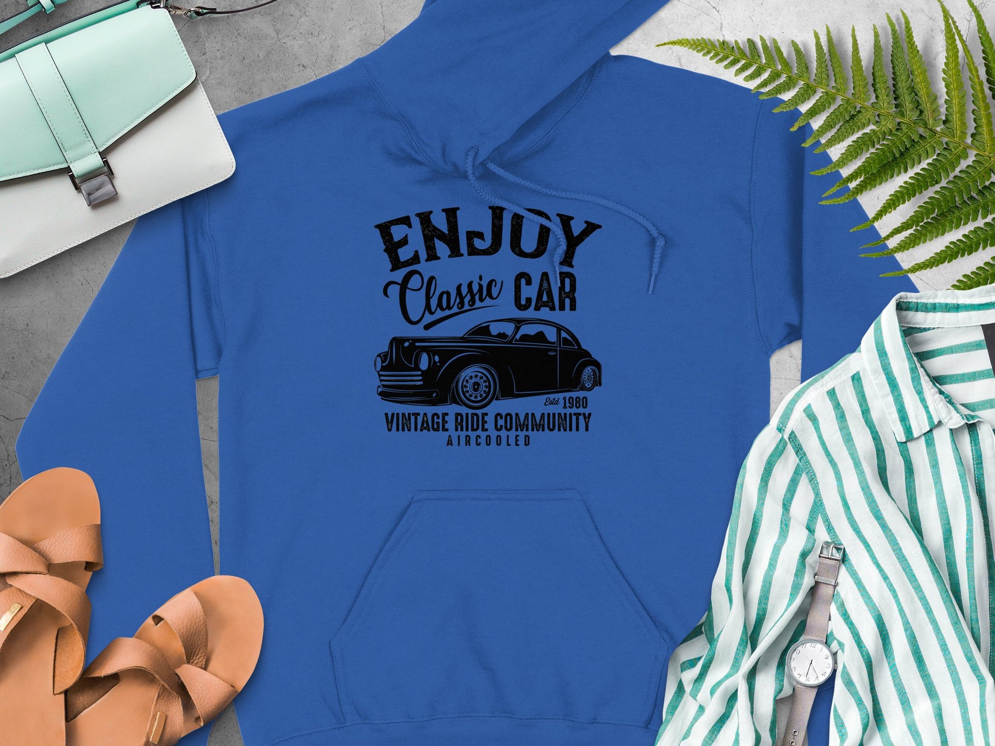 Resting on a gray background with brown sandals, a green and white striped shirt, a wristwatch, a purse, and lush green ferns, this Garment Graphics hoodie features a vintage car graphic with ENJOY Classic CAR, Vintage Ride Community, Aircooled, Estd 1980, exuding true community spirit.