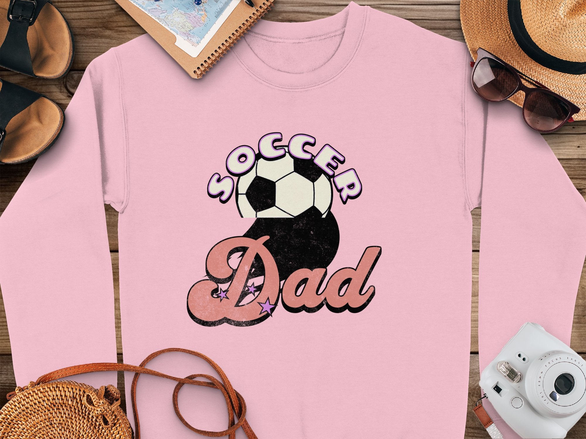 A Garment Graphics retro pink sweatshirt features Soccer Dad with Soccer above a soccer ball graphic and Dad in script font below. Its displayed on a wooden surface with accessories.