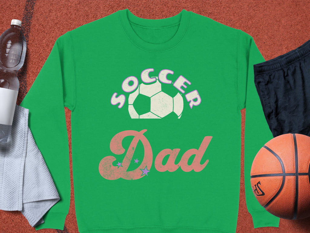 The Garment Graphics Soccer Dad sweatshirt in vibrant green with a soccer ball graphic is showcased with a water bottle, towel, black shorts, and an orange basketball. This unisex sweatshirt perfectly fits any parent cheering on the sidelines.