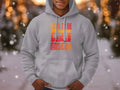 A person showcases a stylish light gray hoodie from Garment Graphics, featuring a vibrant Born To Be Musician design. Blurred bokeh lights and snowy trees highlight the trendy unisex appeal of this cozy attire, perfect for music lovers.