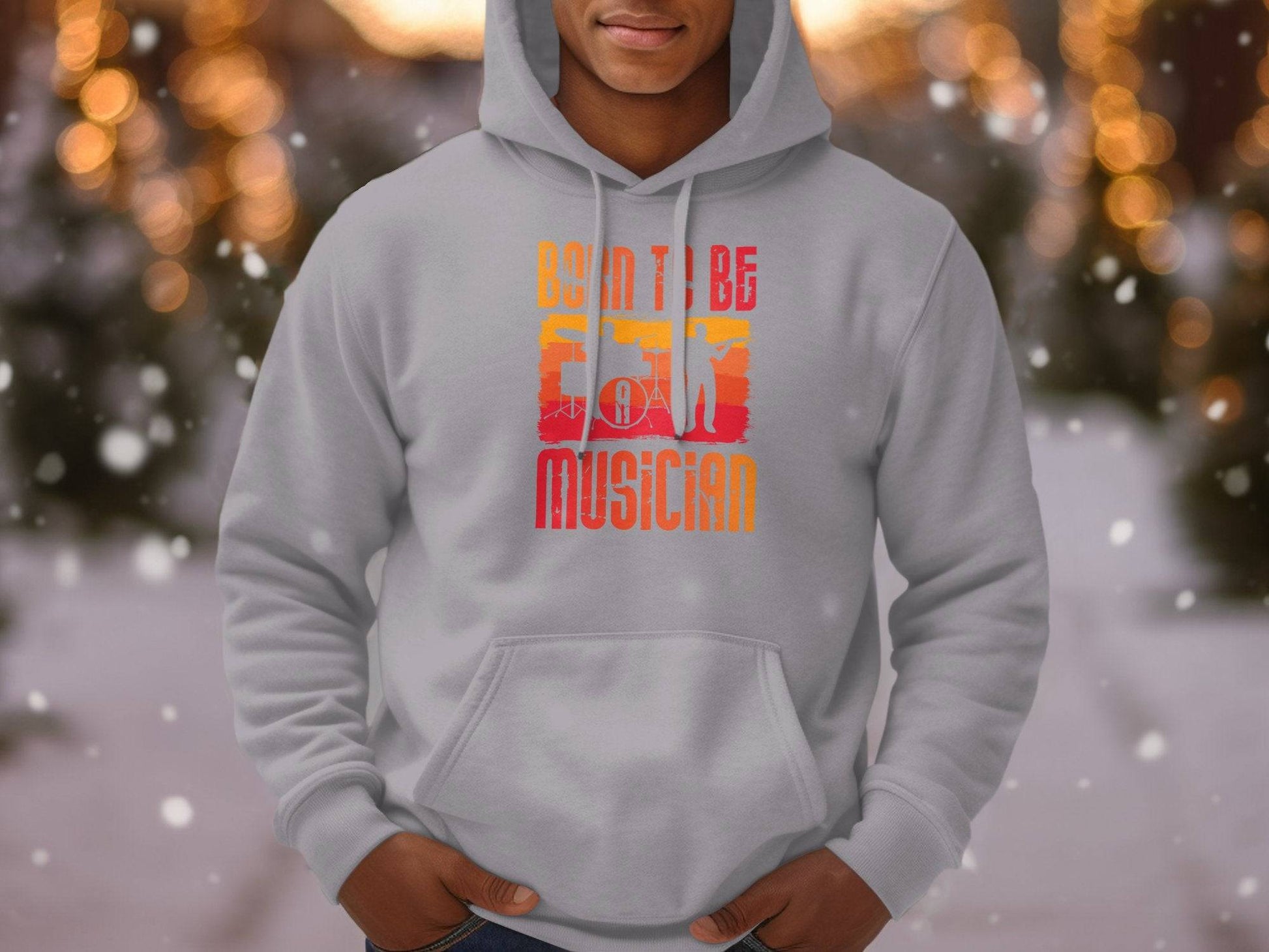 A person showcases a stylish light gray hoodie from Garment Graphics, featuring a vibrant Born To Be Musician design. Blurred bokeh lights and snowy trees highlight the trendy unisex appeal of this cozy attire, perfect for music lovers.