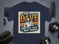 Garment Graphics classic fit navy blue T-shirt showcases a man at a table with the phrase Just Dave Doing Dave Things, surrounded by a smartphone, smartwatch, portable speaker, sneakers, and dumbbells on a gray surface.