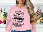 A long-haired blonde woman stands in a cozy kitchen adorned with holiday decor, wearing Garment Graphics pink sweatshirt featuring a vintage car design, celebrating classic car culture reminiscent of Classic London City charm.