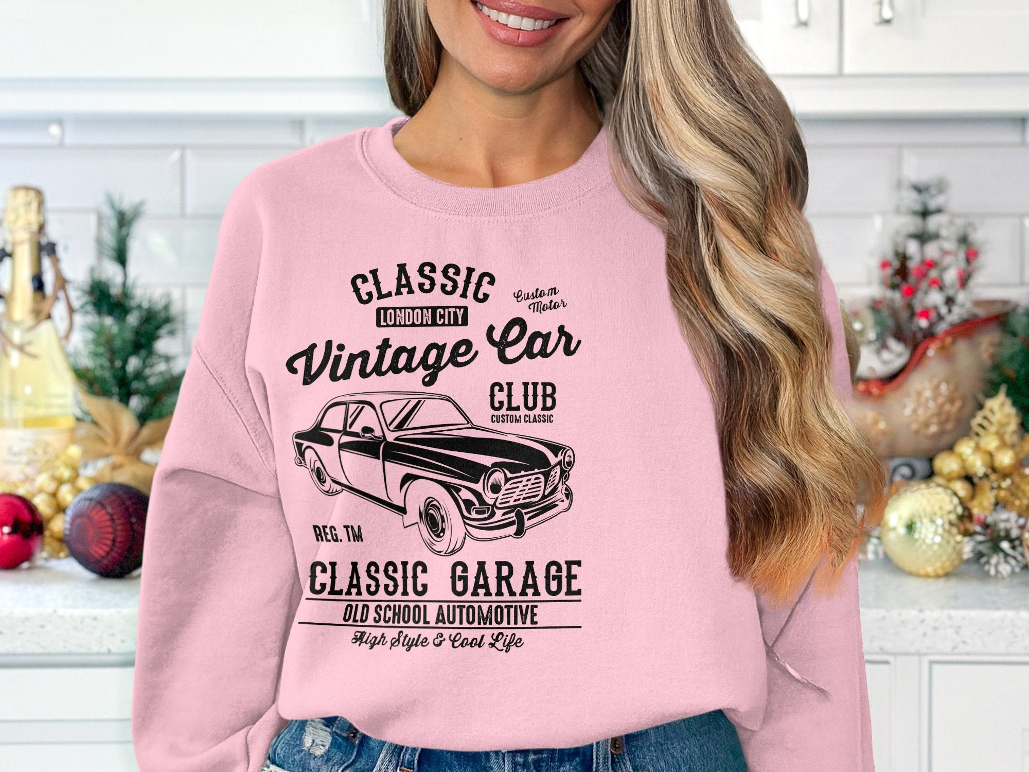 A long-haired blonde woman stands in a cozy kitchen adorned with holiday decor, wearing Garment Graphics pink sweatshirt featuring a vintage car design, celebrating classic car culture reminiscent of Classic London City charm.