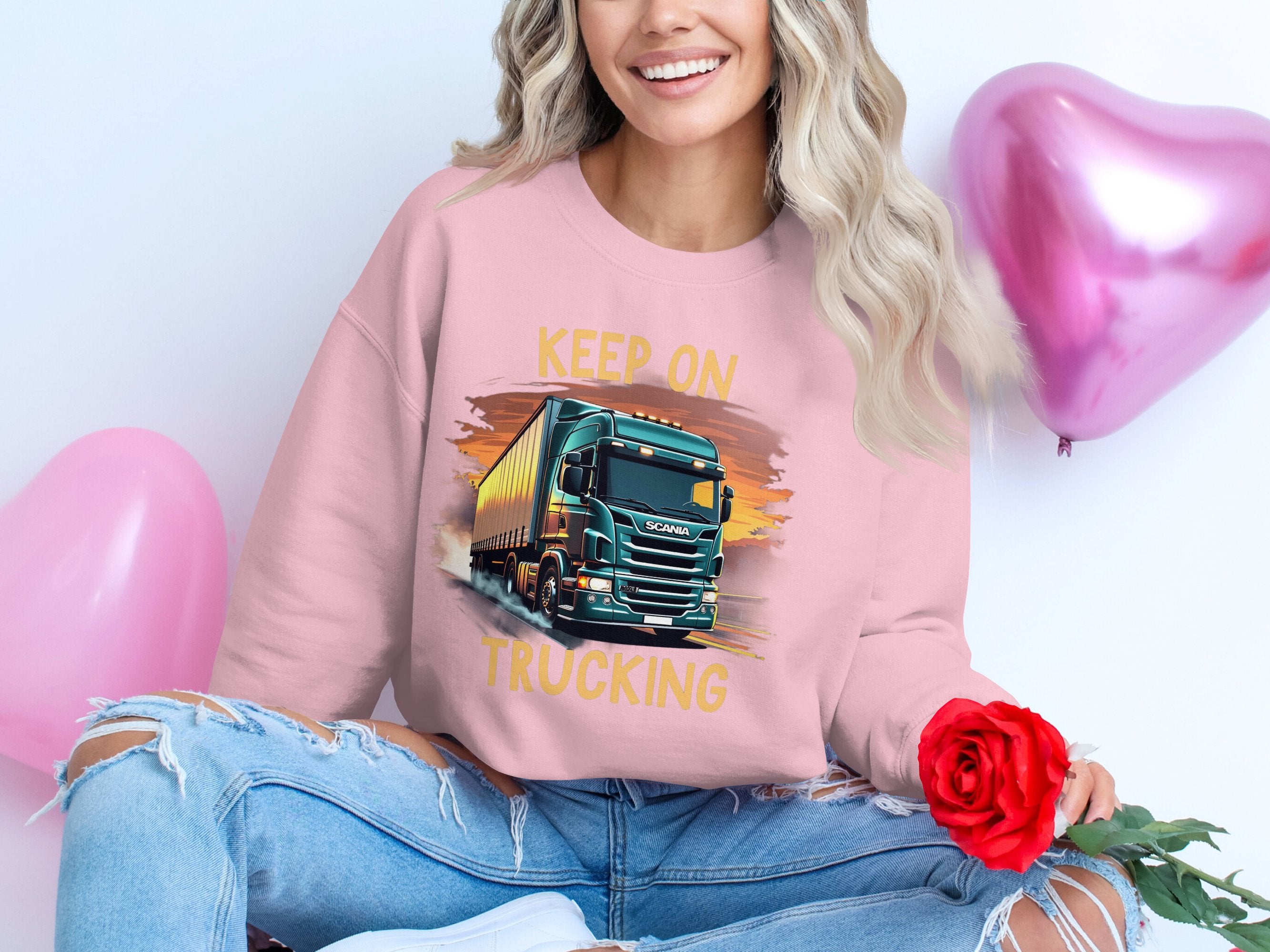A cross-legged person with long, wavy hair wears a Garment Graphics pink Graphic Sweatshirt featuring a truck graphic and Keep On Trucking text. They hold a red rose, while two pink heart-shaped balloons float in the background, ideal for truck enthusiasts.