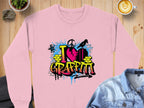 A pink Garment Graphics sweatshirt with a vibrant I Love Graffiti design featuring a heart and arrows is placed on a wooden table. Nearby are a latte, small plant, rock, and partially visible folded denim jacket.