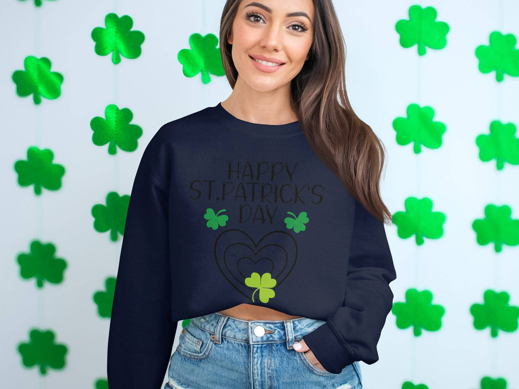 In front of a wall with green shamrock cutouts, a woman radiates warmth in a Garment Graphics sweatshirt featuring Happy St. Patricks Day and clover designs. Her long brown hair falls beautifully as she pairs it with blue denim jeans, smiling cheerfully.