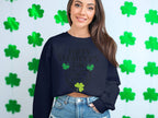 In front of a wall with green shamrock cutouts, a woman radiates warmth in a Garment Graphics sweatshirt featuring Happy St. Patricks Day and clover designs. Her long brown hair falls beautifully as she pairs it with blue denim jeans, smiling cheerfully.
