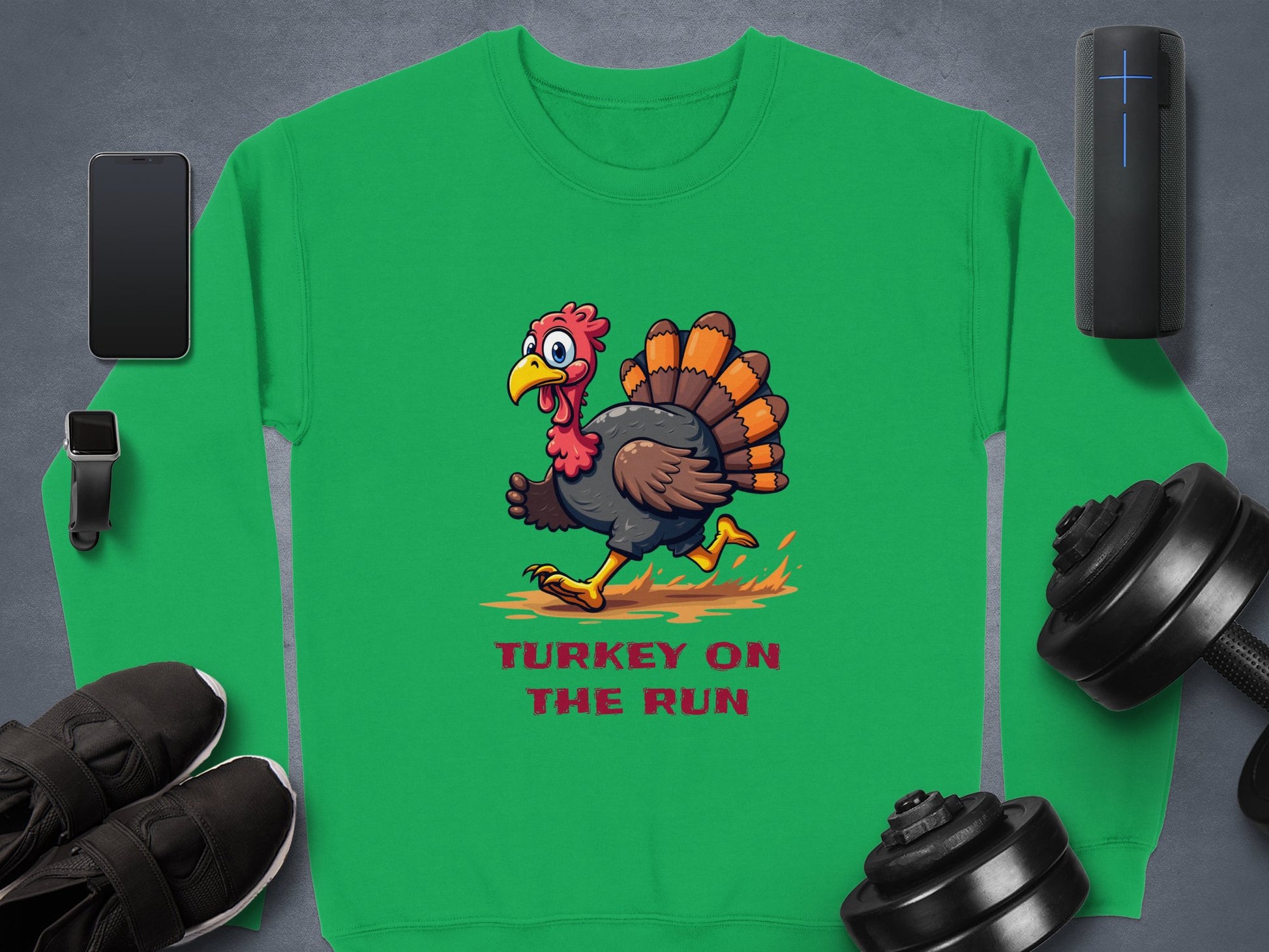 The Garment Graphics unisex green sweatshirt features a cartoon turkey with Turkey on the Run text. It’s styled with a smartphone, smartwatch, black sneakers, weights, and a portable speaker on a gray background.