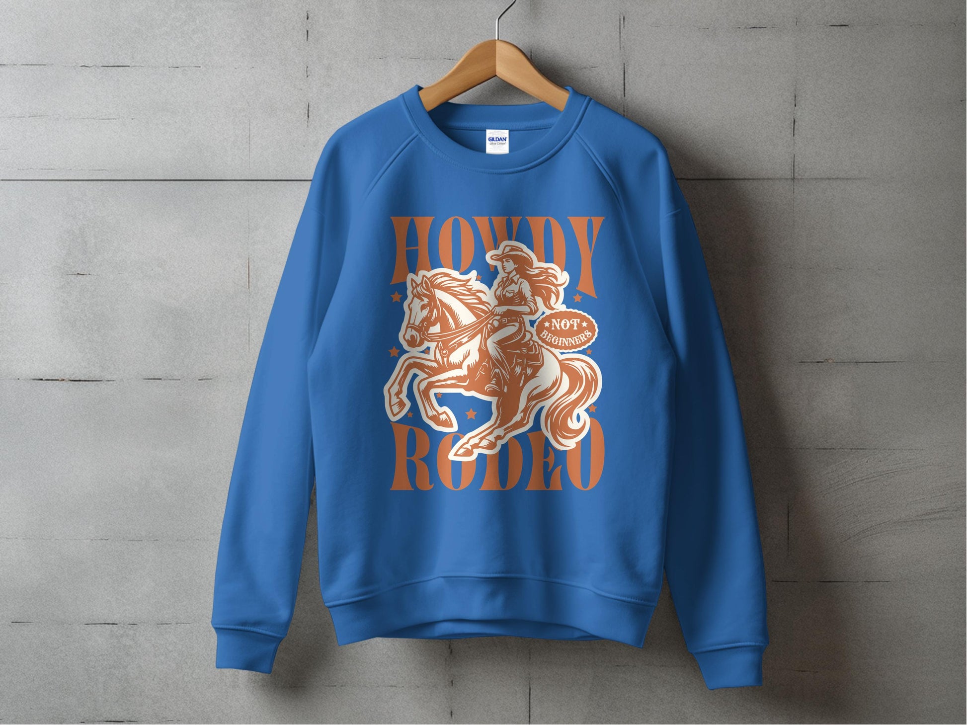 The Garment Graphics blue sweatshirt, perfect for rodeo enthusiasts, is displayed on a gray wall. It features a cowboy riding a horse illustration and the bold Howdy Rodeo text in vibrant orange, capturing the cowboy spirit.