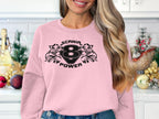In a festive kitchen setting, an individual with long wavy hair and jeans blends seamlessly while donning a Garment Graphics light pink sweatshirt with SCANIR POWER, featuring a stylish print of the number 8 and two bird heads.