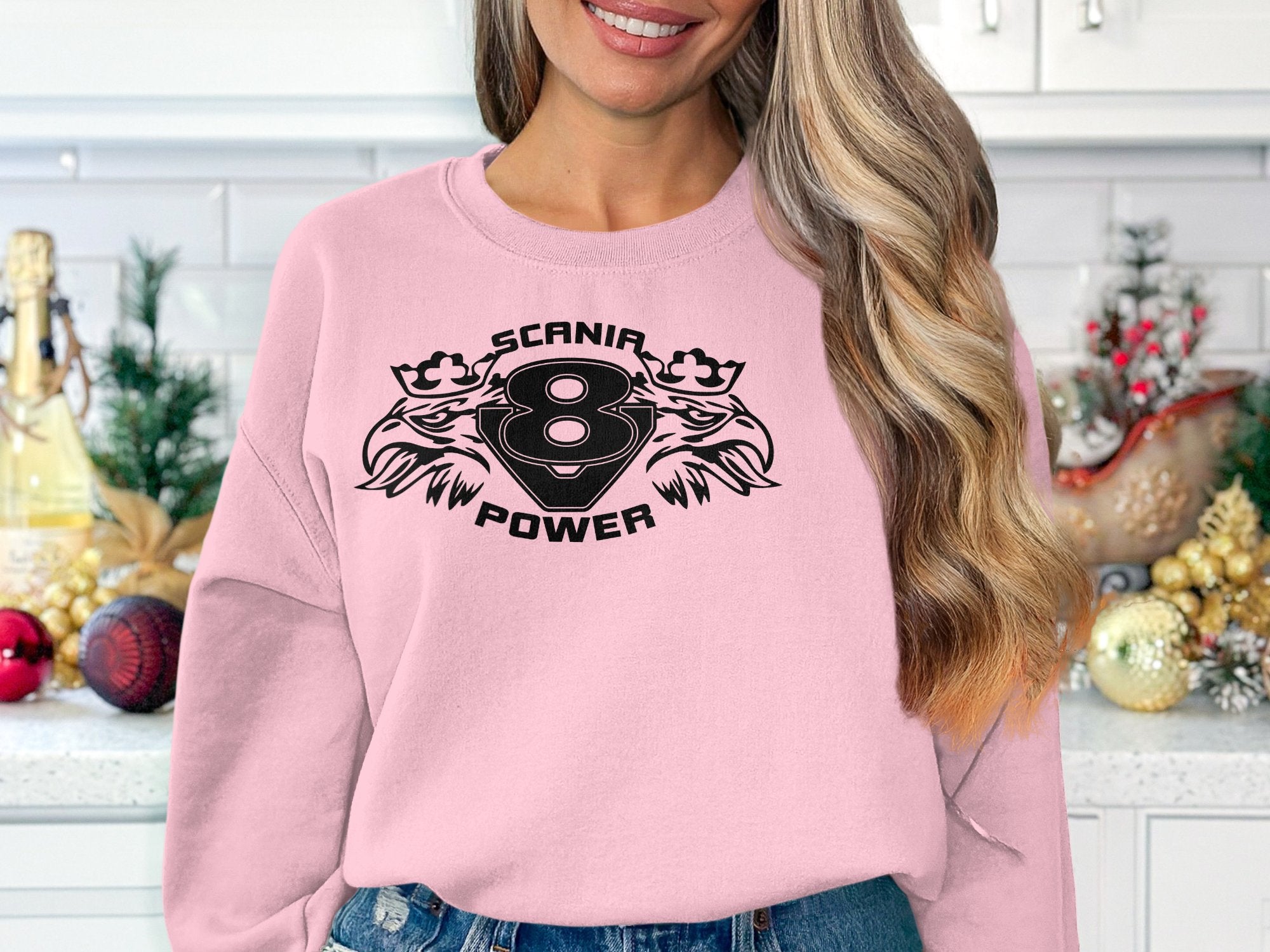 In a festive kitchen setting, an individual with long wavy hair and jeans blends seamlessly while donning a Garment Graphics light pink sweatshirt with SCANIR POWER, featuring a stylish print of the number 8 and two bird heads.