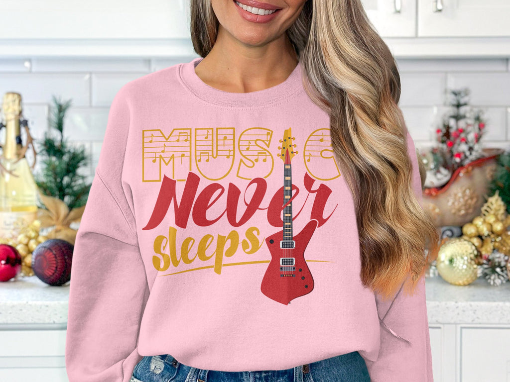 Indoors against a white tiled wall, someone wears Garment Graphics stylish light pink sweatshirt featuring Music Never Sleeps and a vibrant red guitar design, ideal for music enthusiasts. The cozy scene includes a festive countertop with assorted ornaments.