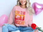 A long-haired person in a Garment Graphics unisex Born to Be Musician pink sweatshirt with a drummer graphic holds a red rose. They wear ripped jeans and are accompanied by two pink heart-shaped balloons, making it an ideal scene for music enthusiasts.