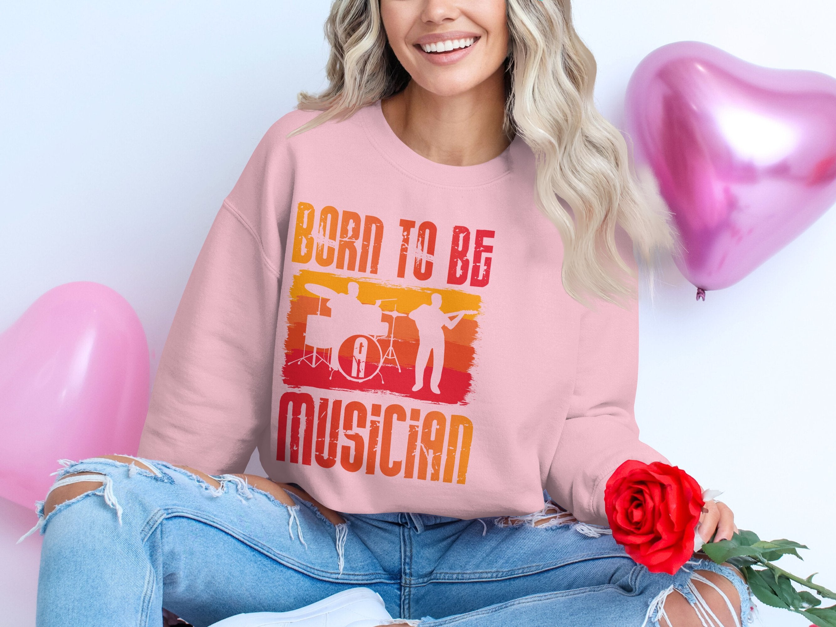 A long-haired person in a Garment Graphics unisex Born to Be Musician pink sweatshirt with a drummer graphic holds a red rose. They wear ripped jeans and are accompanied by two pink heart-shaped balloons, making it an ideal scene for music enthusiasts.