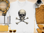 The Garment Graphics white sweatshirt with an edgy skull and crossbones design is placed on a wooden surface. Its complemented by a plaid scarf, quilted jacket, sunglasses, notebook with pen, and coffee mug—all in warm tones that highlight its unisex sizing.