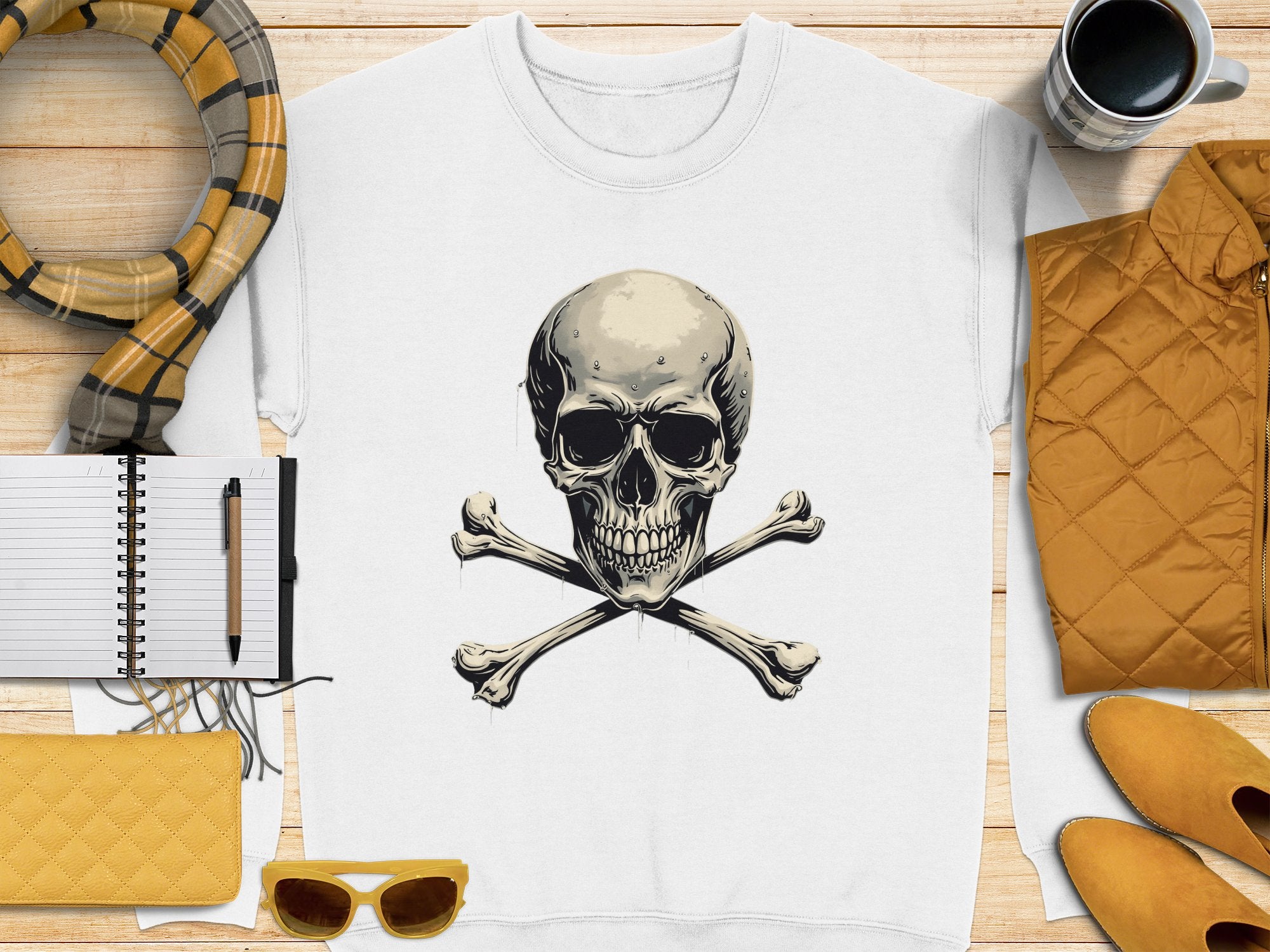 The Garment Graphics white sweatshirt with an edgy skull and crossbones design is placed on a wooden surface. Its complemented by a plaid scarf, quilted jacket, sunglasses, notebook with pen, and coffee mug—all in warm tones that highlight its unisex sizing.