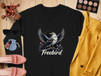 A Garment Graphics black shirt with a Freebird eagle design is paired with a yellow blazer, pink shoes, sunglasses, makeup brushes and palette, and lip products on wood. Its unisex sizing offers wardrobe versatility.