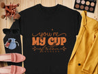 A black Garment Graphics Youre My Cup of Tea unisex tee lies on a wooden surface, encircled by a yellow jacket, sunglasses, makeup brushes, an eyeshadow palette, pink shoes, lipstick, and a small blue pouch.