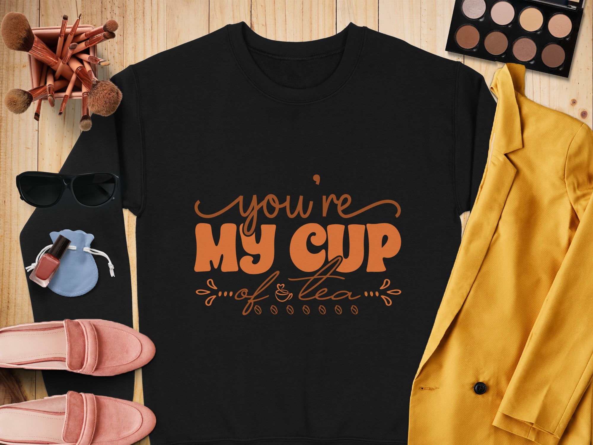 A black Garment Graphics Youre My Cup of Tea unisex tee lies on a wooden surface, encircled by a yellow jacket, sunglasses, makeup brushes, an eyeshadow palette, pink shoes, lipstick, and a small blue pouch.