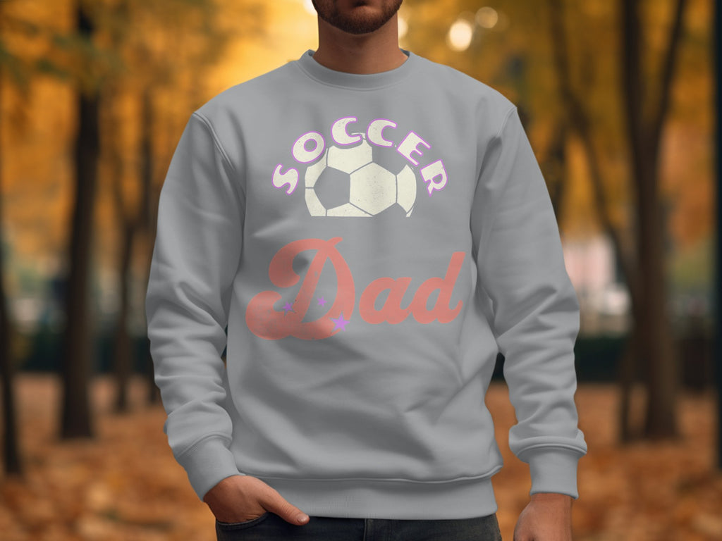 A person stands outdoors in a stylish Soccer Dad sweatshirt from Garment Graphics, featuring a unisex fit and soccer ball graphic. The blurred backdrop showcases trees with vibrant autumn foliage.