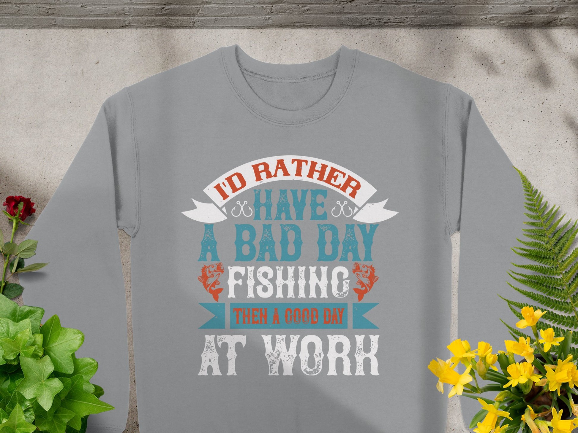 The unisex fishing sweatshirt from Garment Graphics lies surrounded by green and yellow plants. It features the text, Id rather have a bad day fishing than a good day at work, in vibrant blue, orange, and white hues, capturing the essence of passion for fishing apparel.