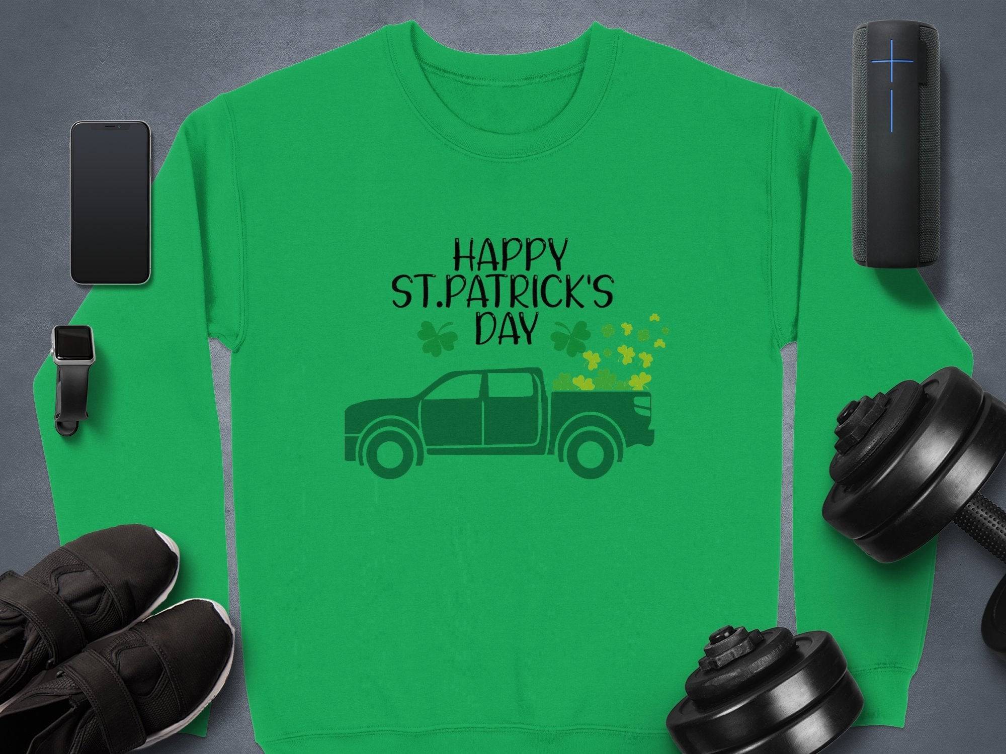 Garment Graphics festive green sweatshirt displays Happy St. Patricks Day with a pickup truck and shamrocks, surrounded by a smartphone, smartwatch, sneakers, dumbbells, Bluetooth speaker, and a cylindrical gadget on a gray surface.