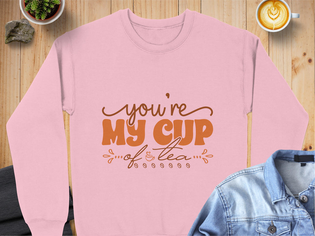 A pink Garment Graphics sweatshirt for tea lovers rests on a wooden surface, emblazoned with youre my cup of tea in brown lettering. Its surrounded by a denim and suit jacket, a latte, a potted plant, and a rock.