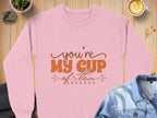 A pink Garment Graphics sweatshirt for tea lovers rests on a wooden surface, emblazoned with youre my cup of tea in brown lettering. Its surrounded by a denim and suit jacket, a latte, a potted plant, and a rock.