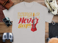 A Garment Graphics 100% cotton gray t-shirt, reading Music Never Sleeps with a red electric guitar graphic, rests on a wooden surface. Nearby are a camera, binoculars, leather pouch, and gloves.