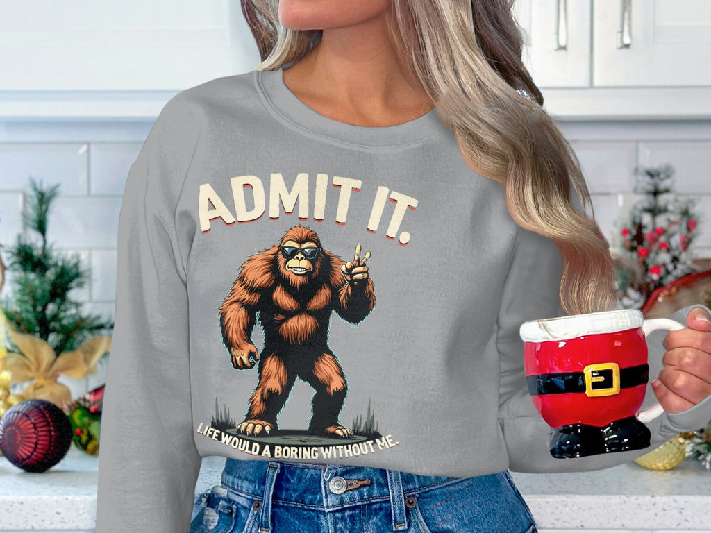 In a festively decorated kitchen, a long-haired person holds a Santa-themed mug while wearing a unisex gray sweatshirt by Garment Graphics that features Bigfoot and the phrase, Admit it, life would be boring without me, offering ultimate comfort.
