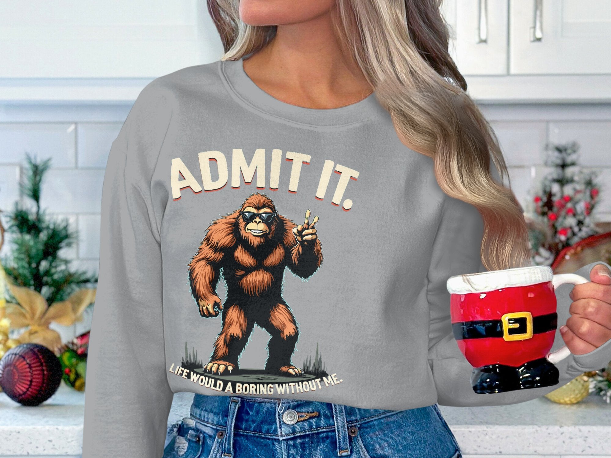 In a festively decorated kitchen, a long-haired person holds a Santa-themed mug while wearing a unisex gray sweatshirt by Garment Graphics that features Bigfoot and the phrase, Admit it, life would be boring without me, offering ultimate comfort.