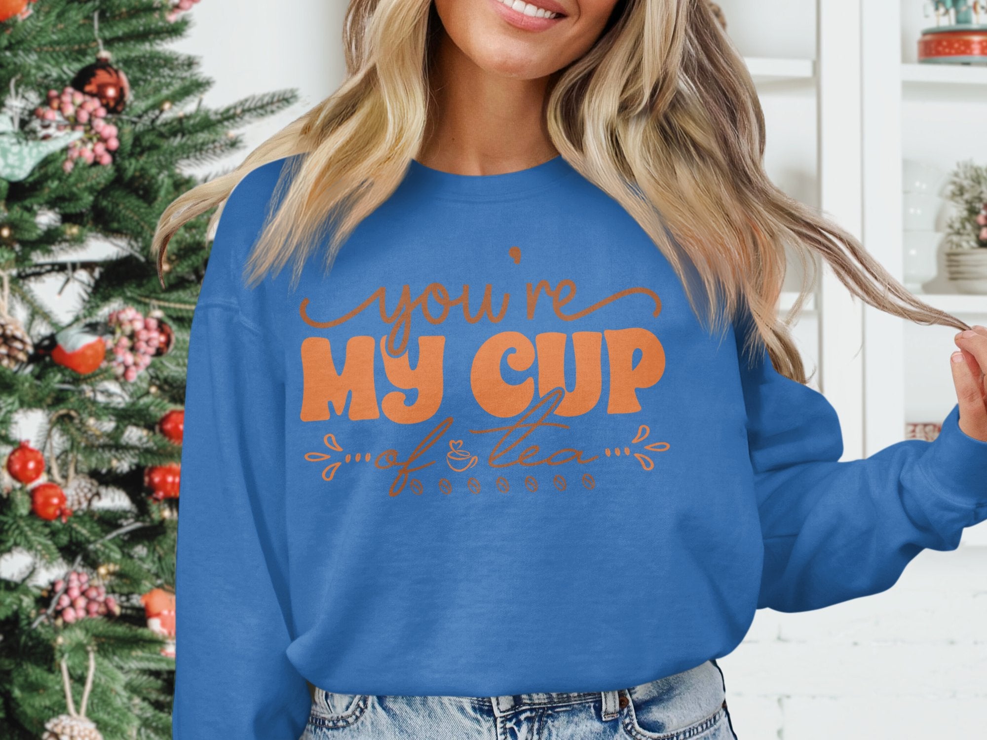 A person wearing a Garment Graphics cozy blue unisex sweatshirt, featuring the orange text youre MY CUP of tea, stands before a decorated Christmas tree and a white cabinet, playfully holding their hair ends.