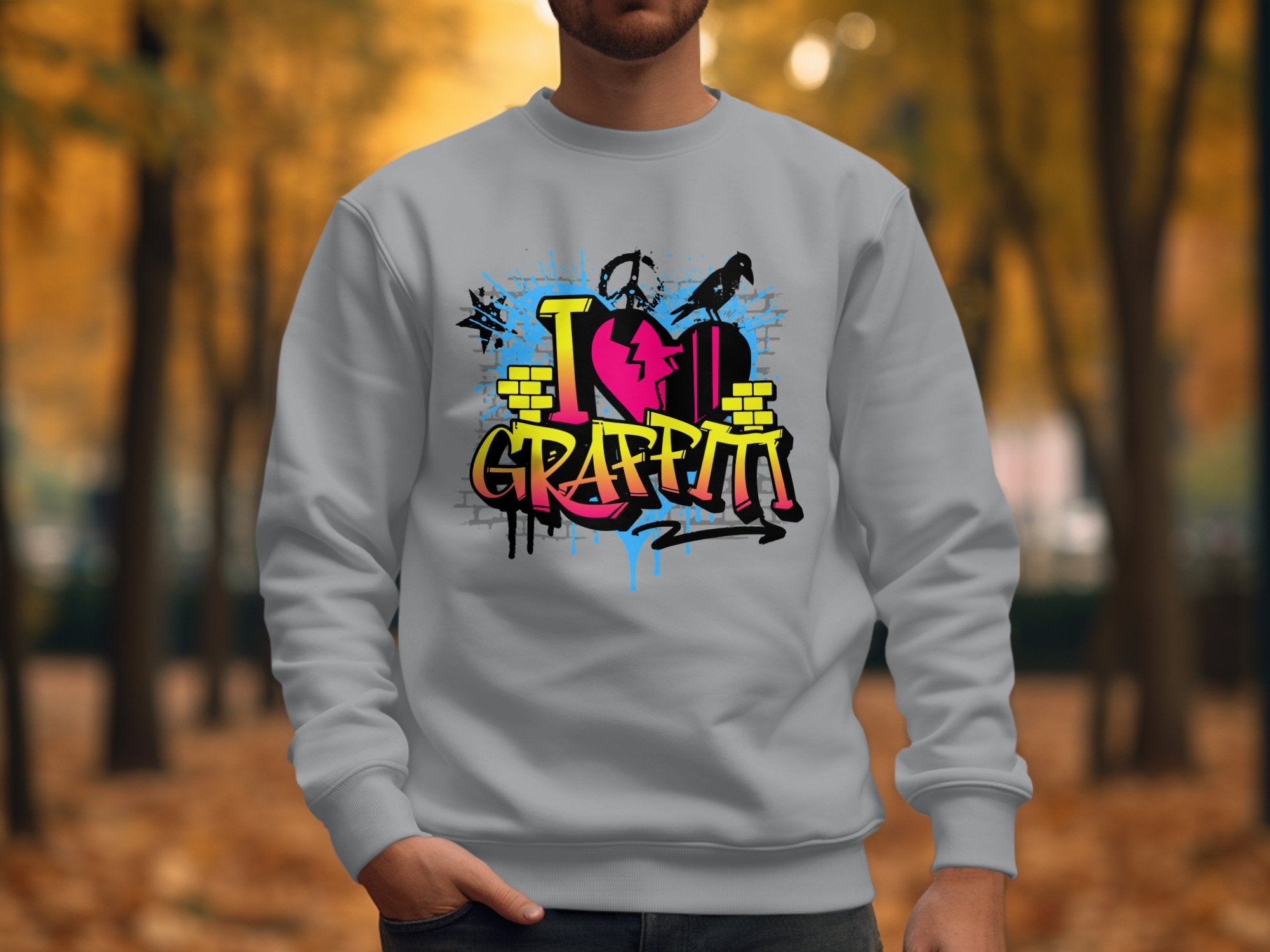 In a park filled with fallen leaves and trees, a person wears a Garment Graphics sweatshirt featuring vibrant graffiti-style art with I Love Graffiti text and a cracked red heart design.
