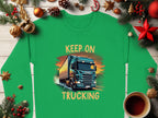 Garment Graphics Graphic Sweatshirt features a bold truck design with KEEP ON TRUCKING above and below, framed by Christmas decorations like ornaments, pinecones, and a cozy coffee cup on a wooden surface.