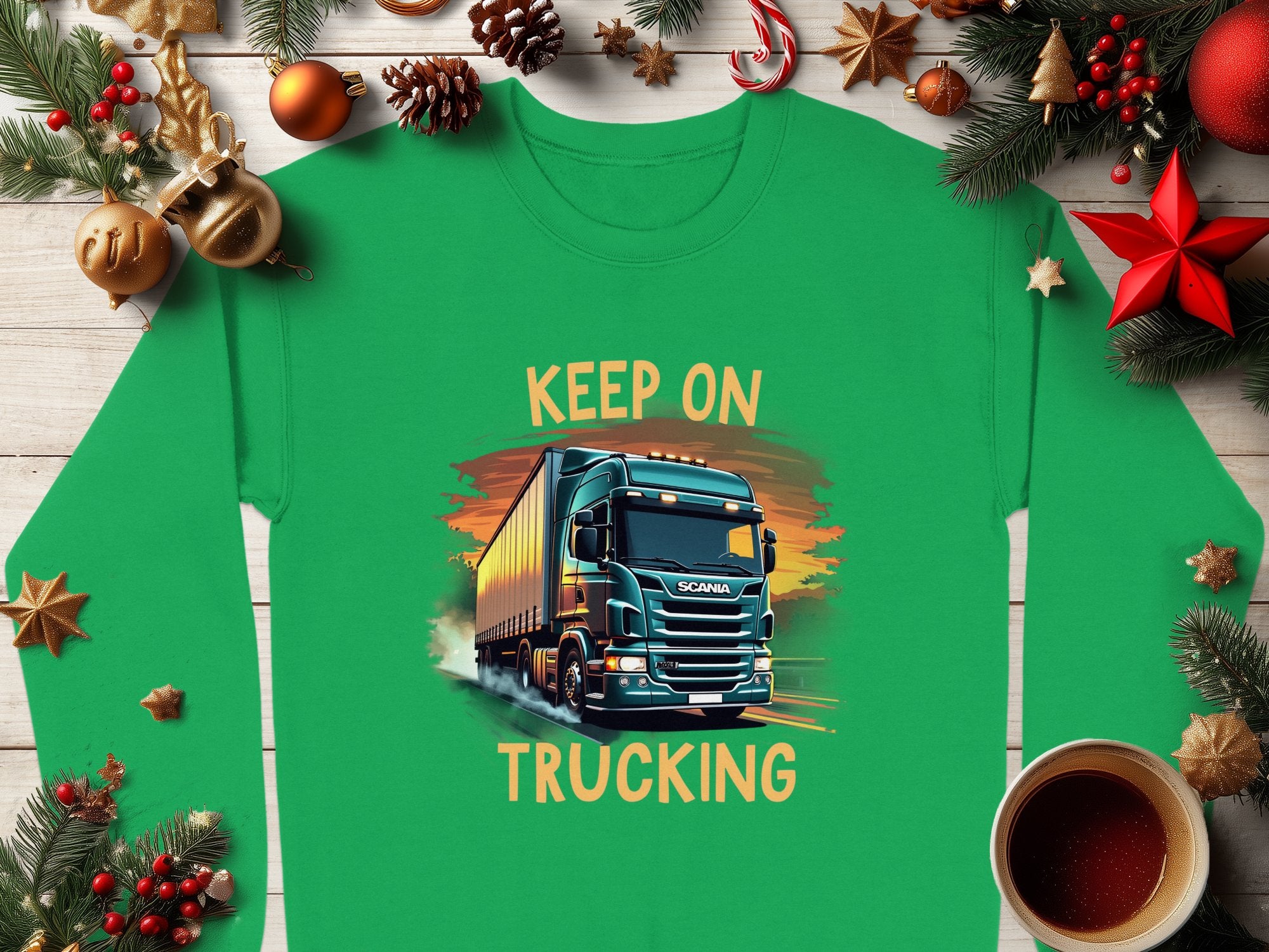 Garment Graphics Graphic Sweatshirt features a bold truck design with KEEP ON TRUCKING above and below, framed by Christmas decorations like ornaments, pinecones, and a cozy coffee cup on a wooden surface.