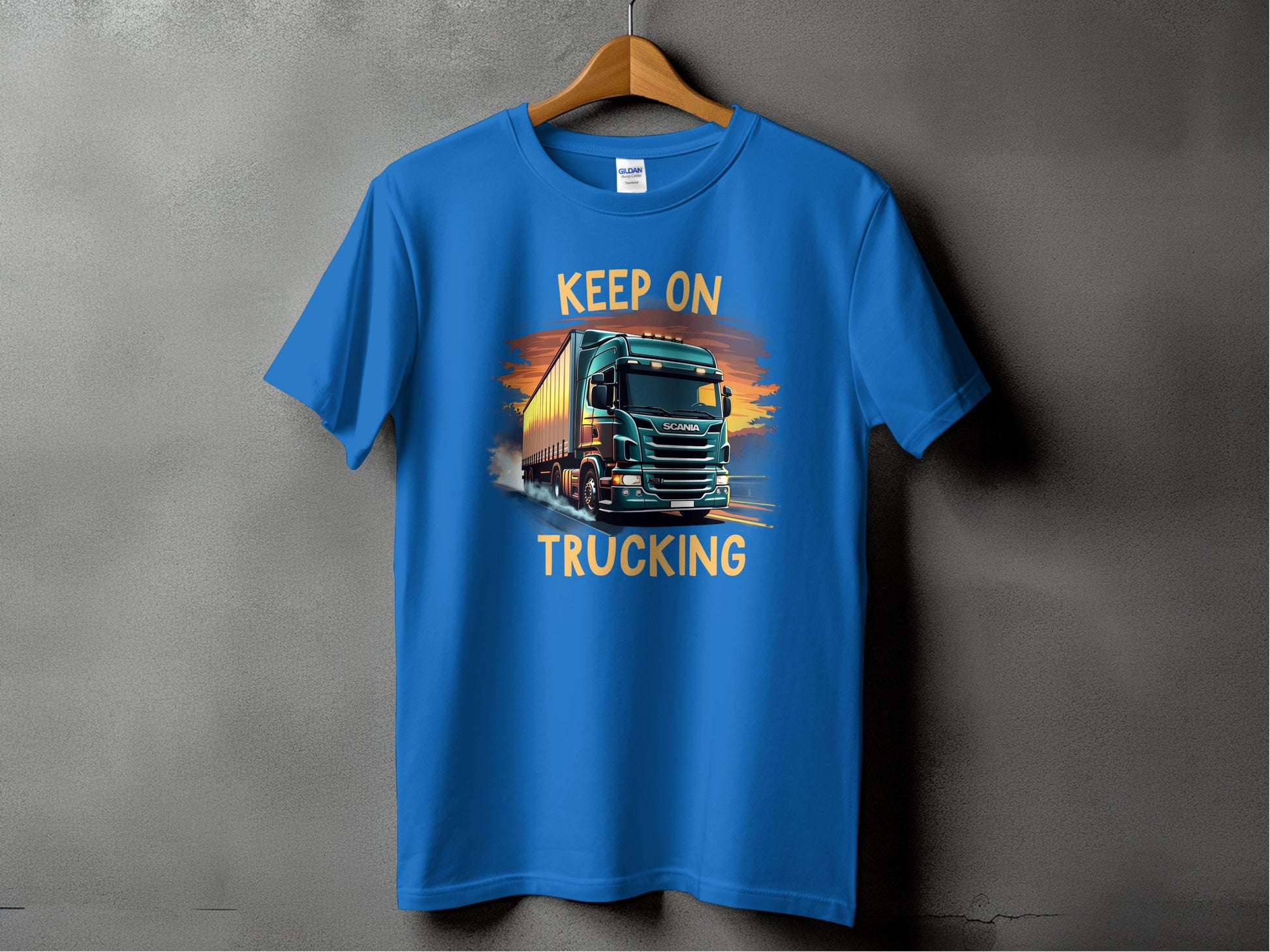 A blue Garment Graphics T-shirt hangs on a wooden hanger against a gray backdrop, ideal for truck fans. It features a semi-truck illustration with the bold text Keep On Trucking above and below the vehicle.