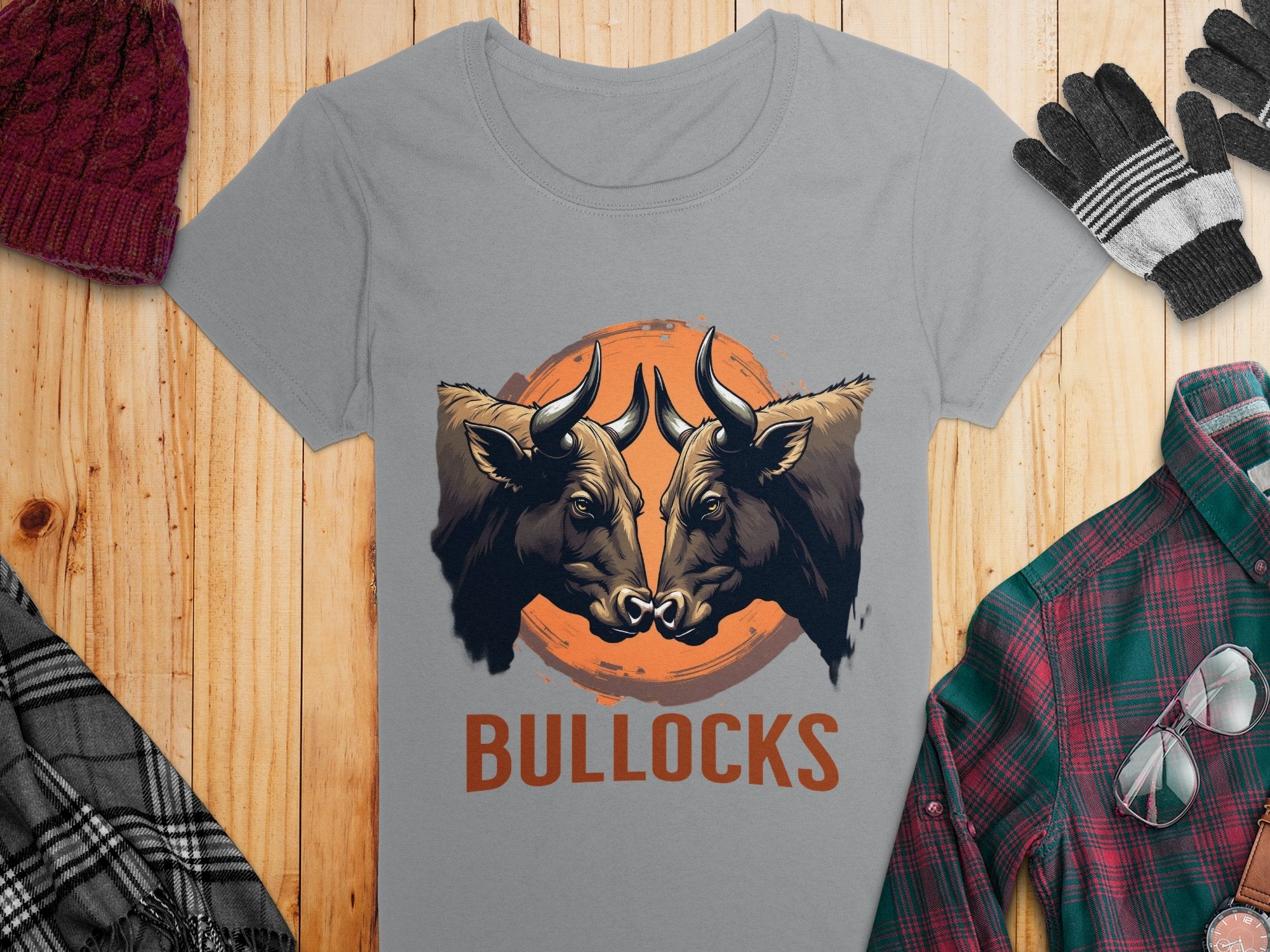 A gray Garment Graphics t-shirt showcases bold graphics of two bull heads on an orange circle with BULLOCKS printed below. Styled casually with a beanie, gloves, and plaid shirt on a wooden surface.