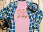 A pink T-shirt from Garment Graphics with This Day is Powered by Coffee sits on a wooden surface, radiating positivity. Its paired with a green and blue plaid shirt for layering, alongside headphones, a camera, and a disposable coffee cup.