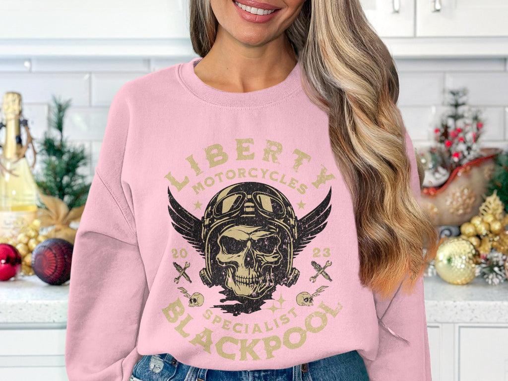 A woman in a light pink sweatshirt by Garment Graphics, showcasing a biker design with a skull, wings, goggles, and Liberty Motorcycles Specialist Blackpool 2023, stands in a festively decorated kitchen.