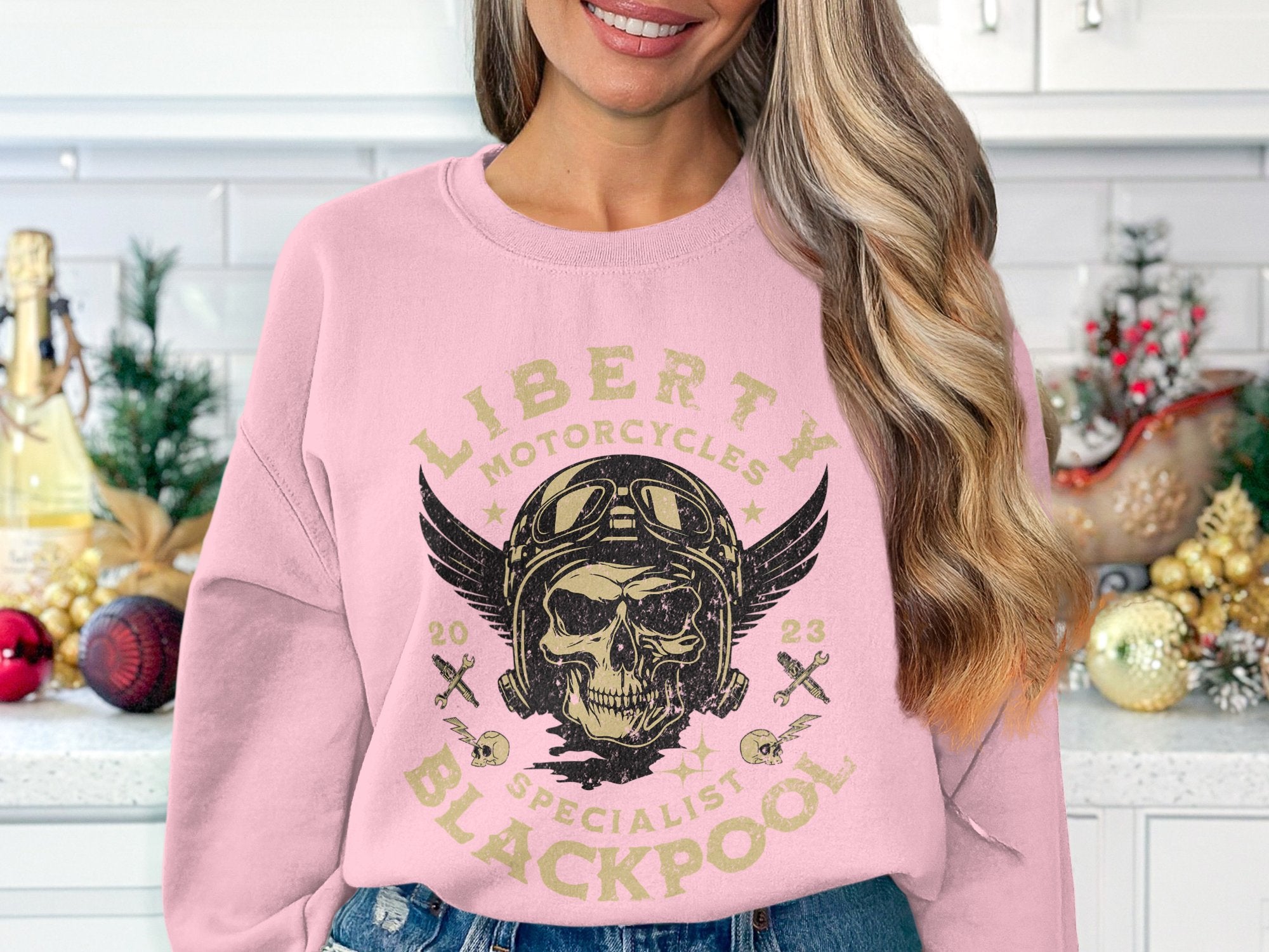A woman in a light pink sweatshirt by Garment Graphics, showcasing a biker design with a skull, wings, goggles, and Liberty Motorcycles Specialist Blackpool 2023, stands in a festively decorated kitchen.