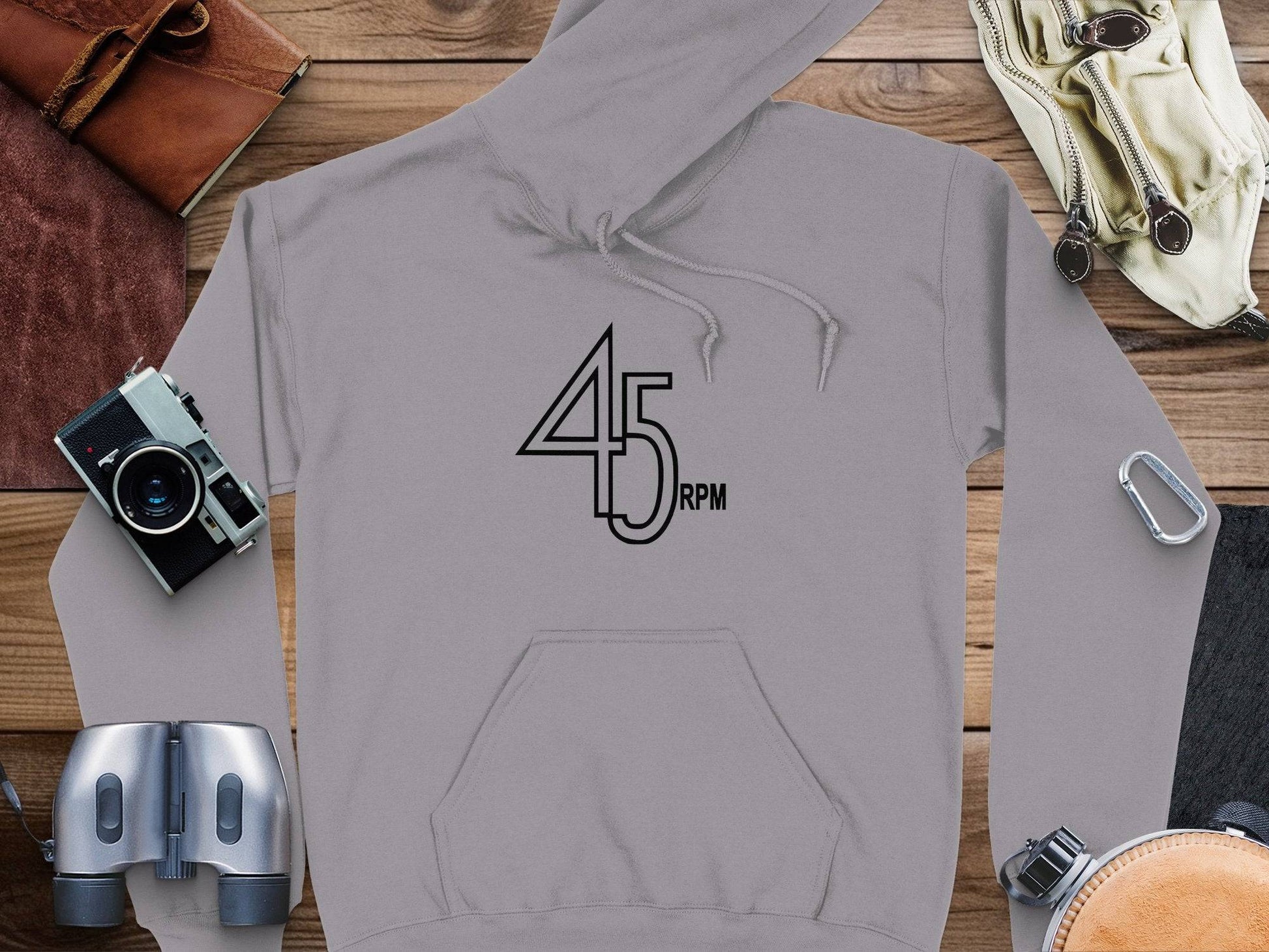 Garment Graphics presents a gray hoodie featuring a 45 RPM design on wood. Its surrounded by items like a camera, binoculars, and a brown wallet, making it perfect for vinyl enthusiasts and fans of retro music-themed graphic hoodies.