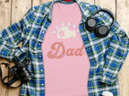 A pink Soccer Dad T-shirt from Garment Graphics is styled with a blue and green plaid shirt, alongside black headphones, a camera, a coffee cup lid, and a phone on a wooden surface.