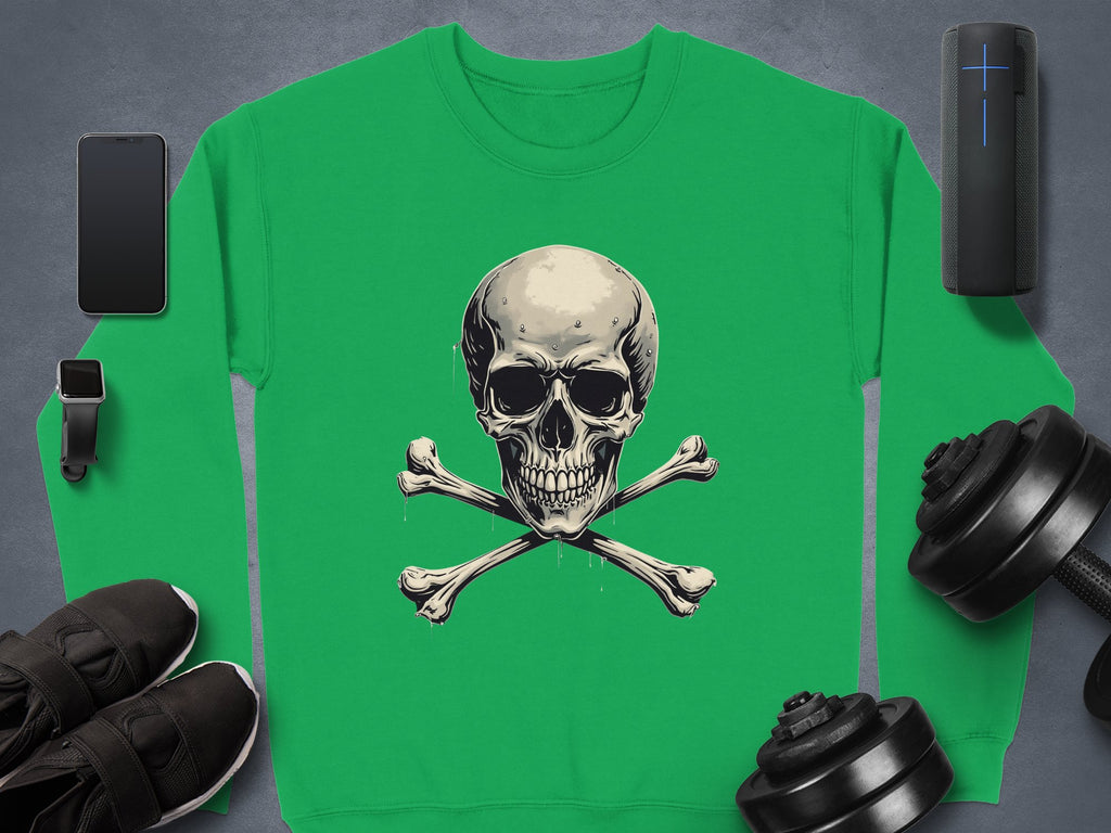 A green Garment Graphics sweater with a skull and crossbones graphic lies on a gray surface, embodying edgy style. Nearby are a smartphone, smartwatch, black sneakers, portable speaker, and dumbbells—items enhancing its unisex sizing allure.