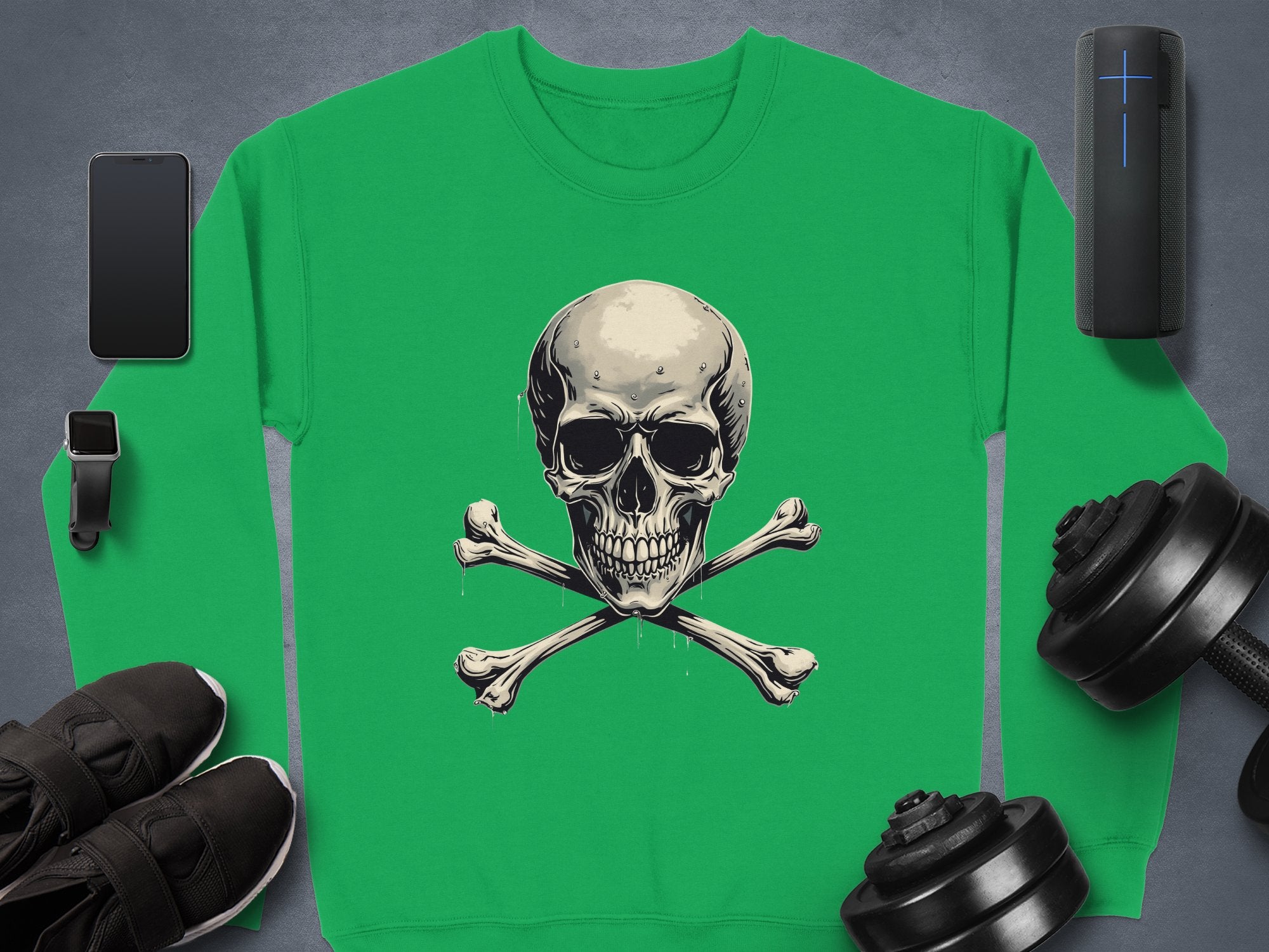A green Garment Graphics sweater with a skull and crossbones graphic lies on a gray surface, embodying edgy style. Nearby are a smartphone, smartwatch, black sneakers, portable speaker, and dumbbells—items enhancing its unisex sizing allure.
