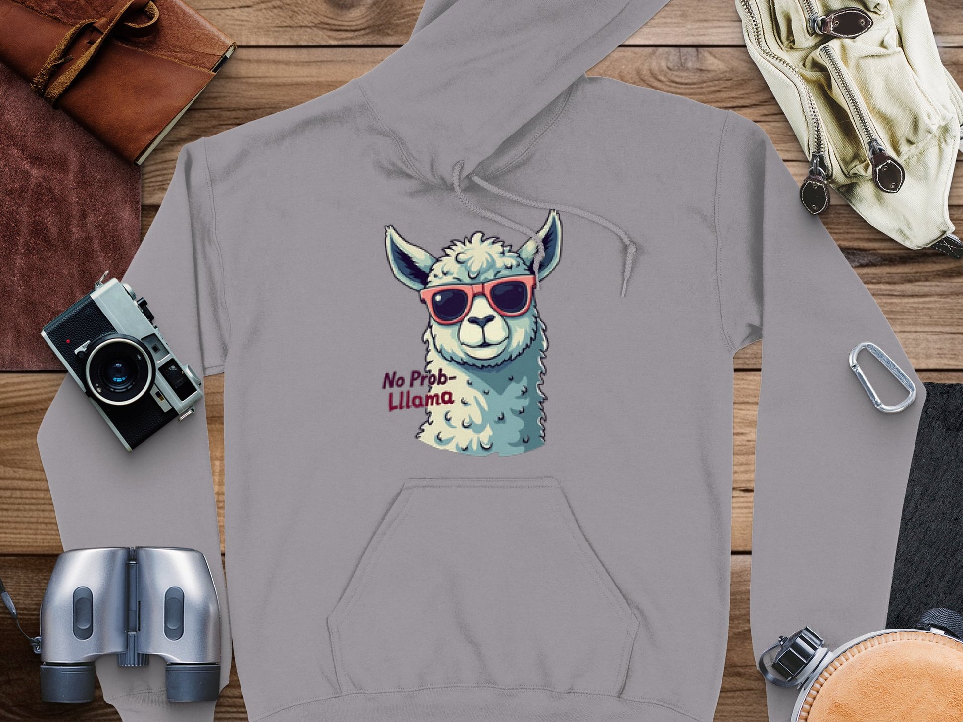 The Garment Graphics unisex grey hoodie showcases a llama in red sunglasses with No Prob-Llama text, surrounded artfully by a vintage camera, binoculars, brown leather item, notepad, and gloves on a wooden surface.