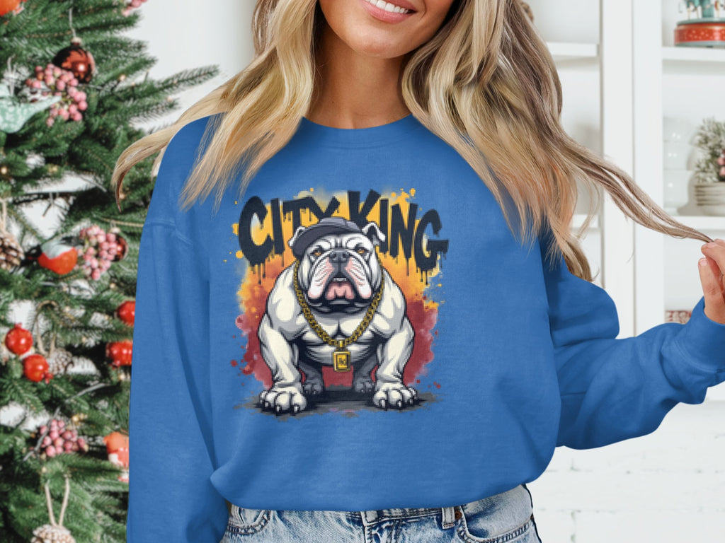Wearing a Garment Graphics Bulldog Sweatshirt in blue, featuring a bulldog graphic with a cap and gold chain and surrounded by CITY KING, a person stands before a decorated Christmas tree.