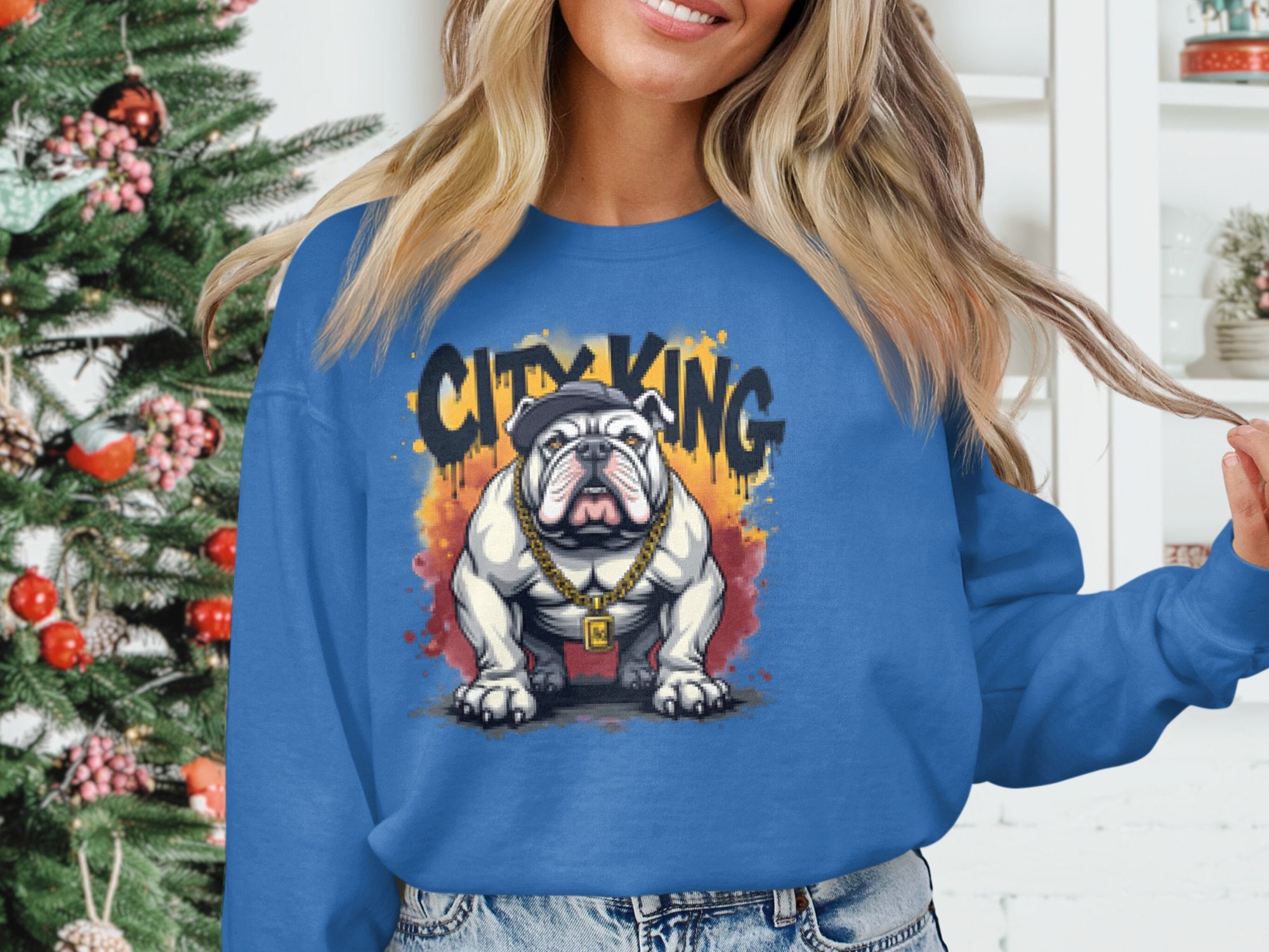 Wearing a Garment Graphics Bulldog Sweatshirt in blue, featuring a bulldog graphic with a cap and gold chain and surrounded by CITY KING, a person stands before a decorated Christmas tree.