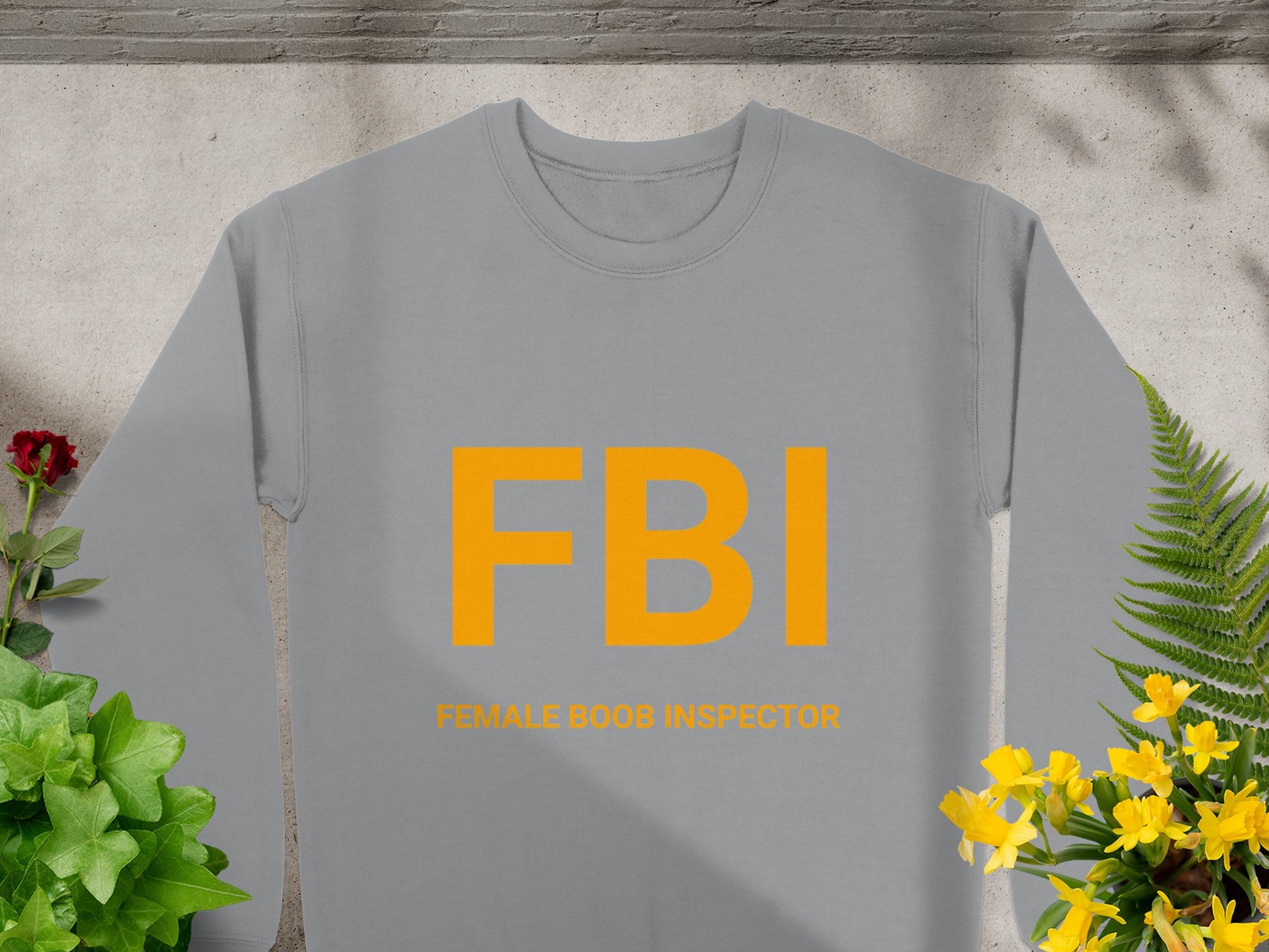 A gray sweatshirt by Garment Graphics displays a FBI graphic in bold yellow above Female Boob Inspector. Unisex sizing, it is showcased on concrete, surrounded by green plants and yellow flowers.