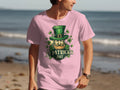 A person strolls along the beach wearing a pink St. Patricks Day T-shirt by Garment Graphics, featuring an Irish pride design with a green-hatted leprechaun and shamrocks, set against the ocean backdrop.